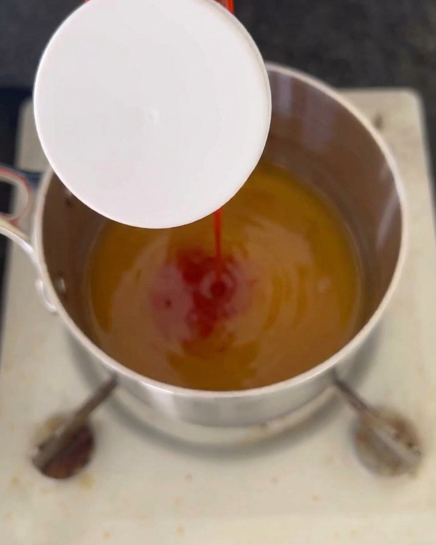 Un chorro de ketchup rojo vertiéndose en una mezcla líquida de color ámbar que burbujea dentro de una olla de acero inoxidable en la estufa.