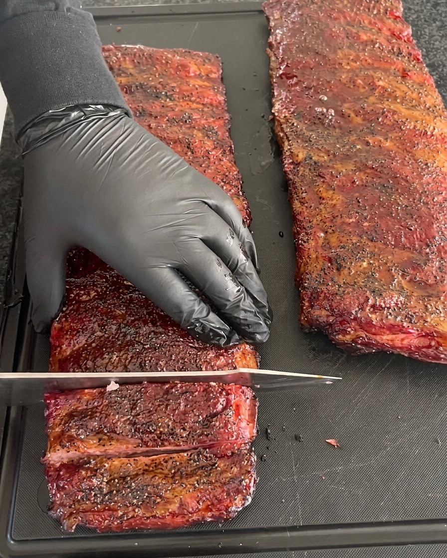 Una persona con guante negro sujetando un costillar ahumado con una corteza profunda mientras corta costillas individuales con un cuchillo grande.