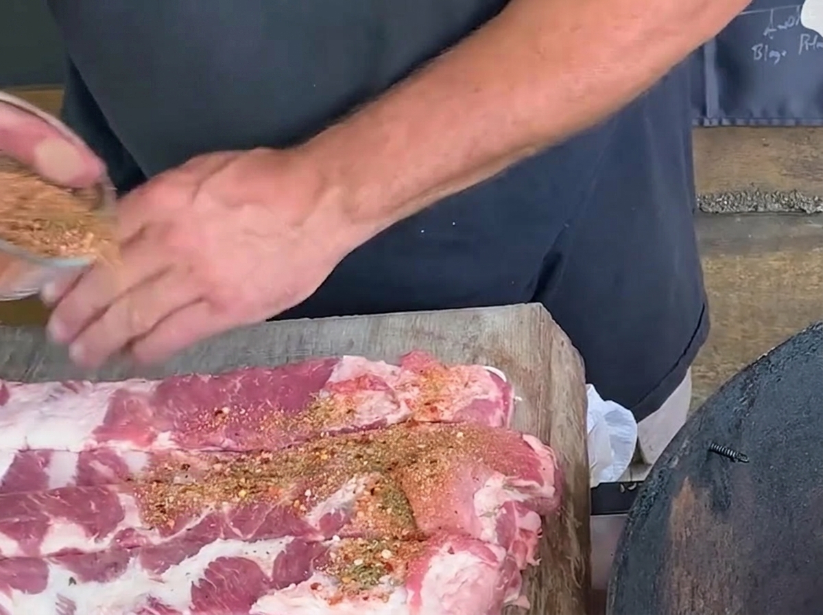 Una persona espolvoreando una mezcla de especias desde un cuenco de vidrio sobre tiras largas de costillas de cerdo crudas en una tabla rústica de madera.