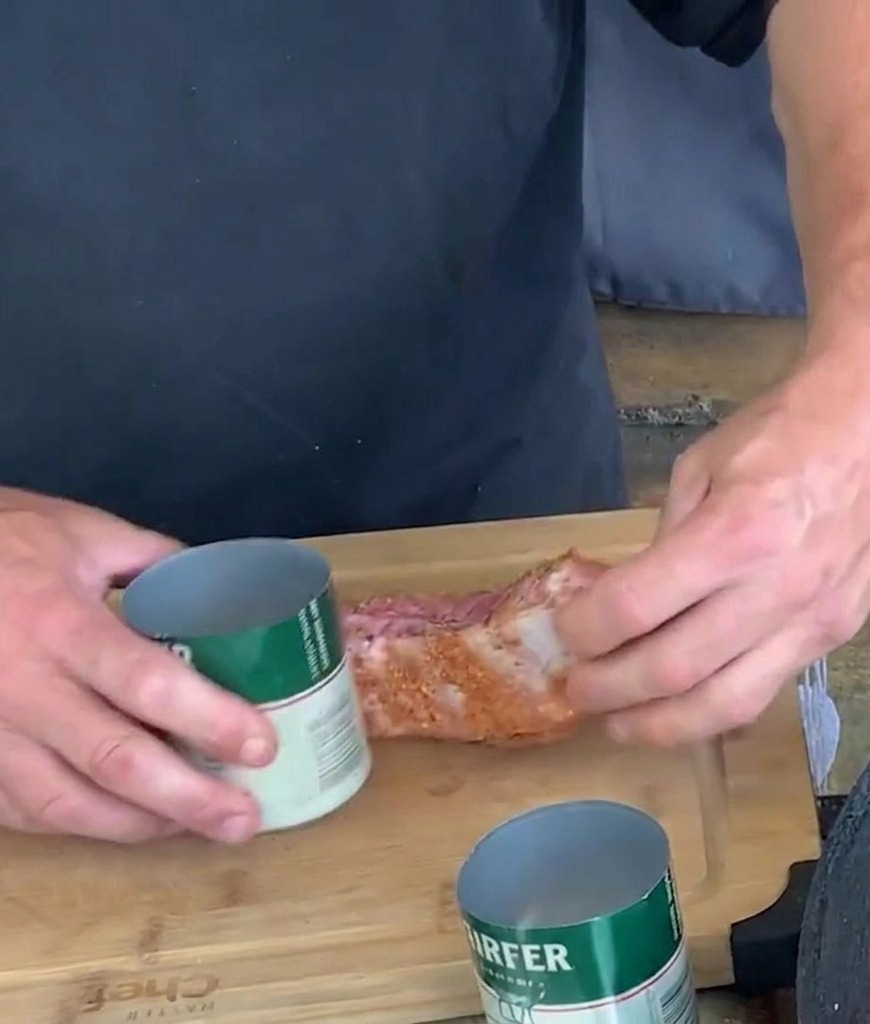 Manos moldeando una tira de costilla de cerdo sazonada envolviéndola alrededor de una lata de aluminio abierta verde y blanca sobre una tabla de cortar.
