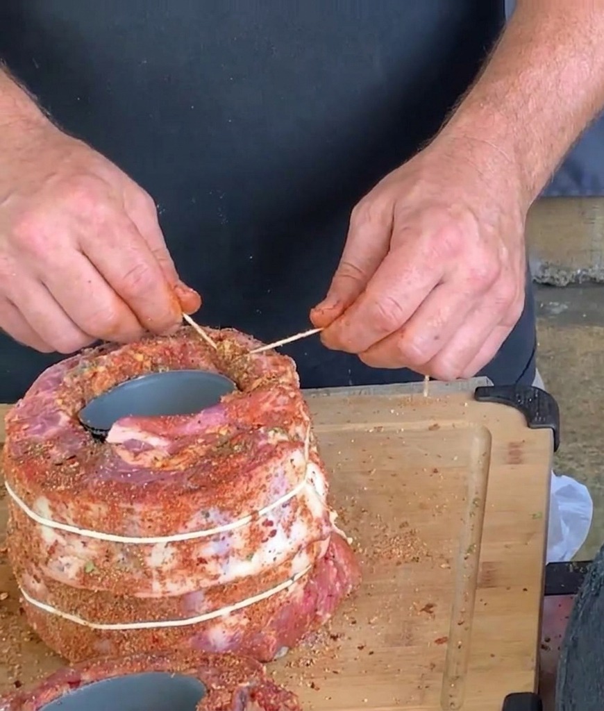 Manos usando hilo blanco de cocina y un palillo para atar y asegurar un gran rollo cilíndrico de costillas de cerdo sazonadas envueltas alrededor de una lata.