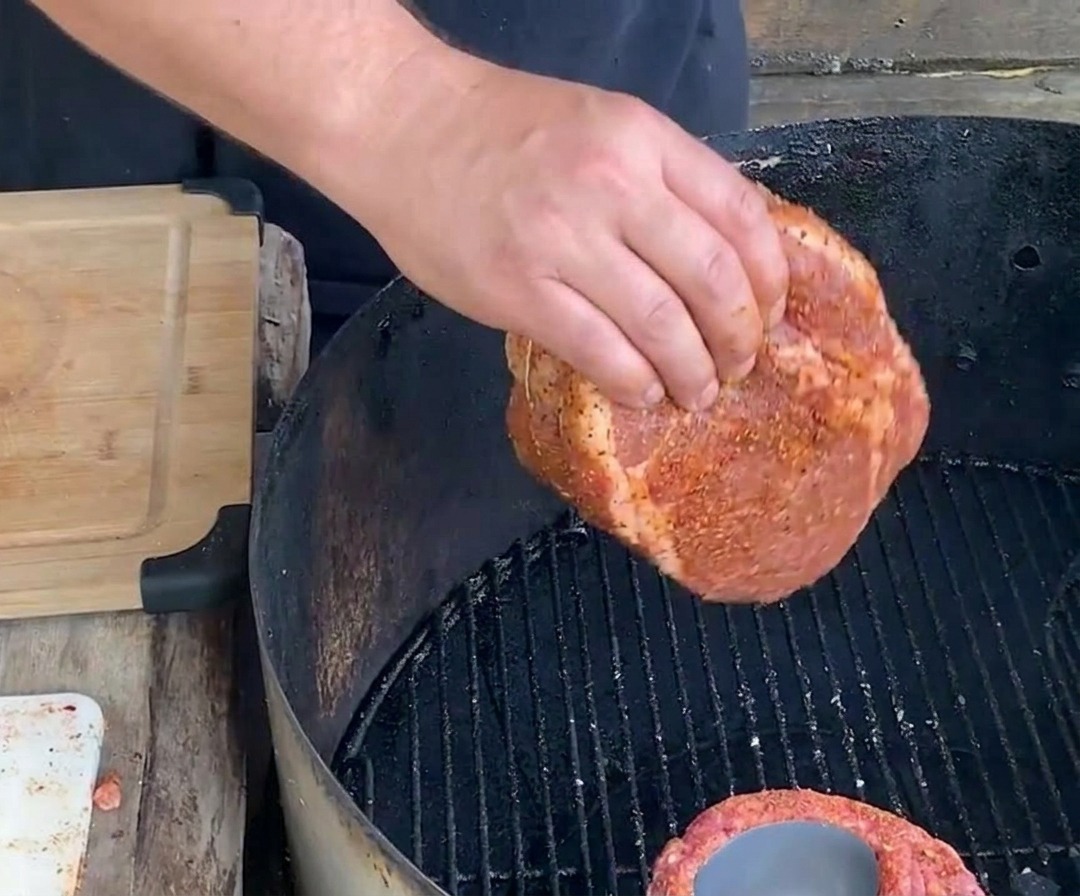Una mano colocando una copa de costilla sazonada y atada, con la lata en su interior, sobre la rejilla negra de una parrilla de carbón para un asado lento.