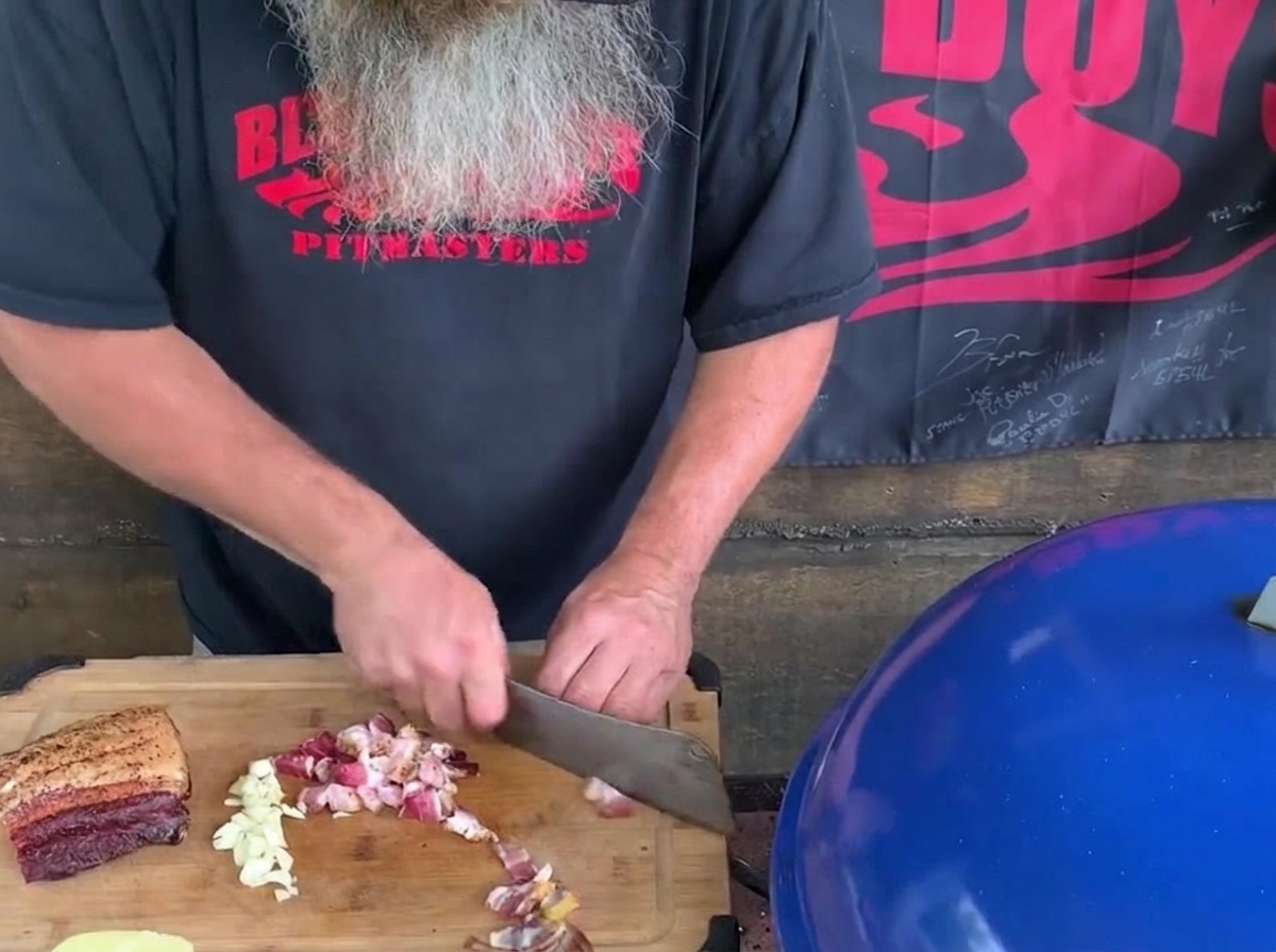 Un hombre con barba larga y gris usando un gran hacha de cocina para picar trozos de bacon y dientes de ajo en una tabla de cortar de madera junto a una parrilla azul.