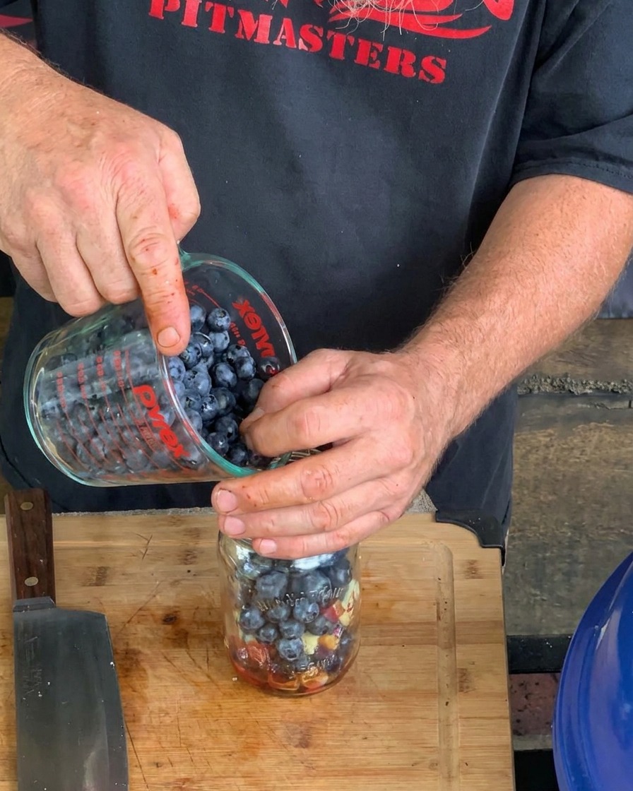 Un plano detalle de manos vertiendo arándanos frescos desde una taza medidora Pyrex en un frasco de vidrio que contiene otros ingredientes líquidos y sólidos para la salsa.
