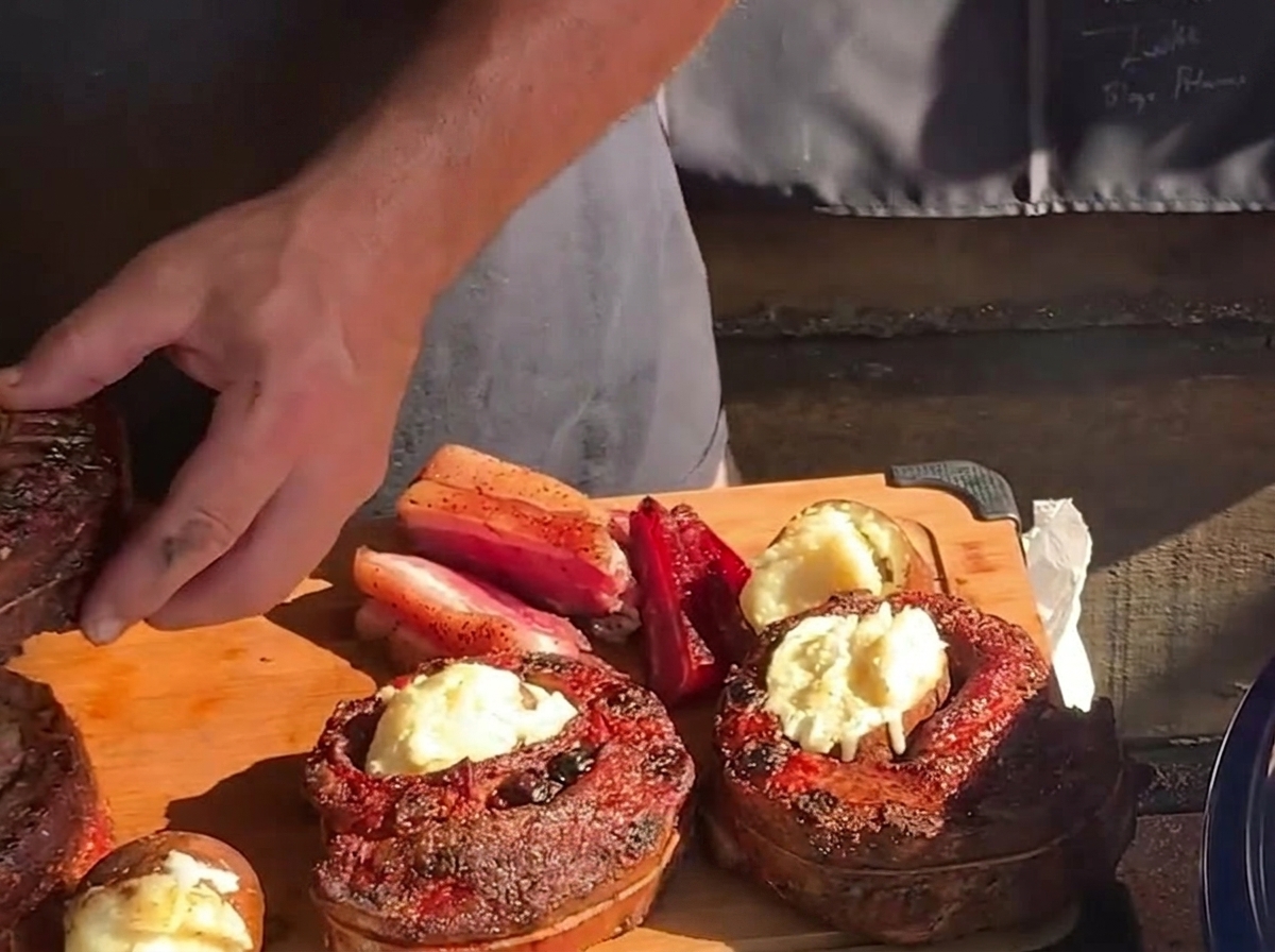 Una mano introduciendo una patata asada caliente con mantequilla directamente en el centro hueco de una copa de costilla ya cocinada sobre una tabla de madera.