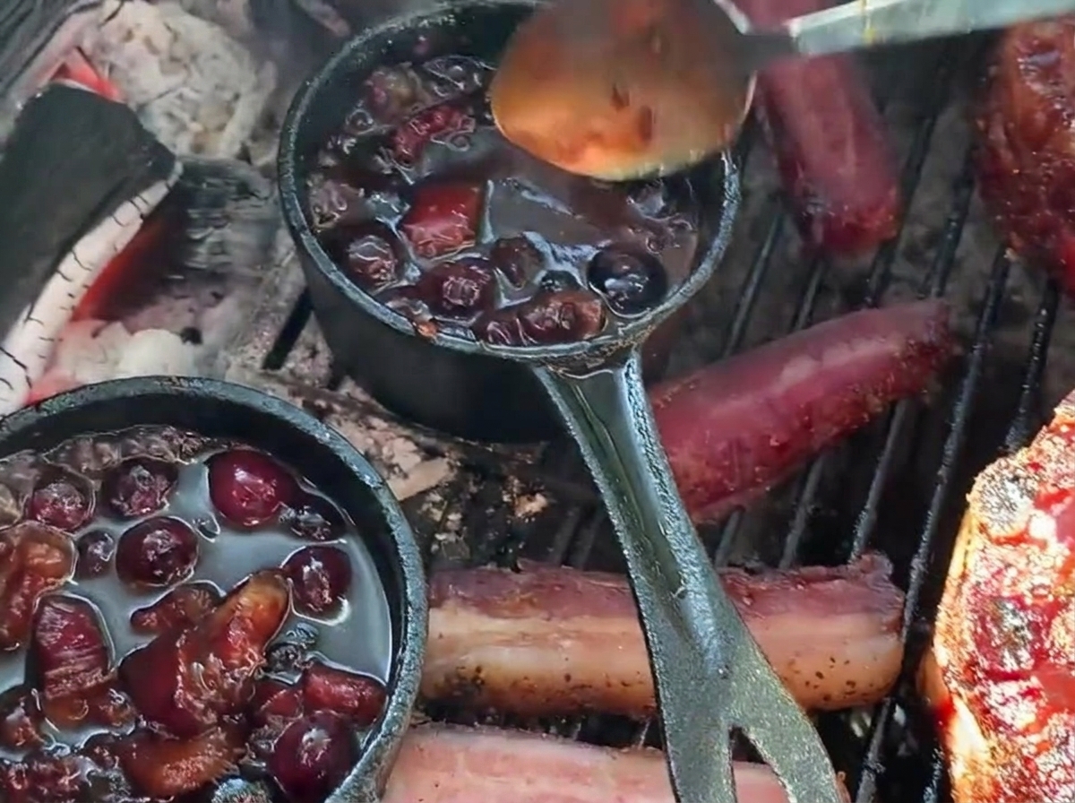 Vertiendo con una cuchara una rica salsa barbacoa de arándanos desde una pequeña sartén de hierro fundido sobre una copa de costilla rellena en la parrilla.