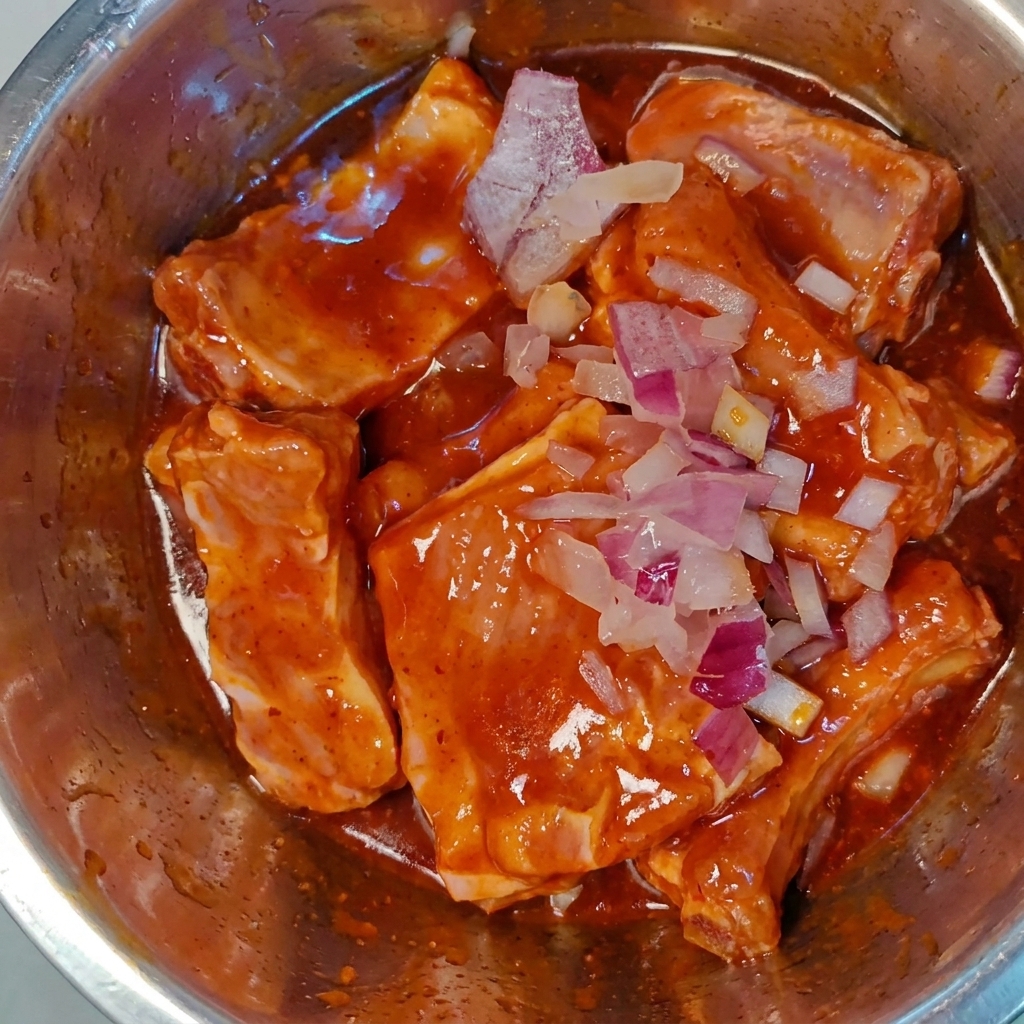 Cebolla morada finamente picada esparcida sobre costillas de cerdo crudas maridadas en un bol de metal.