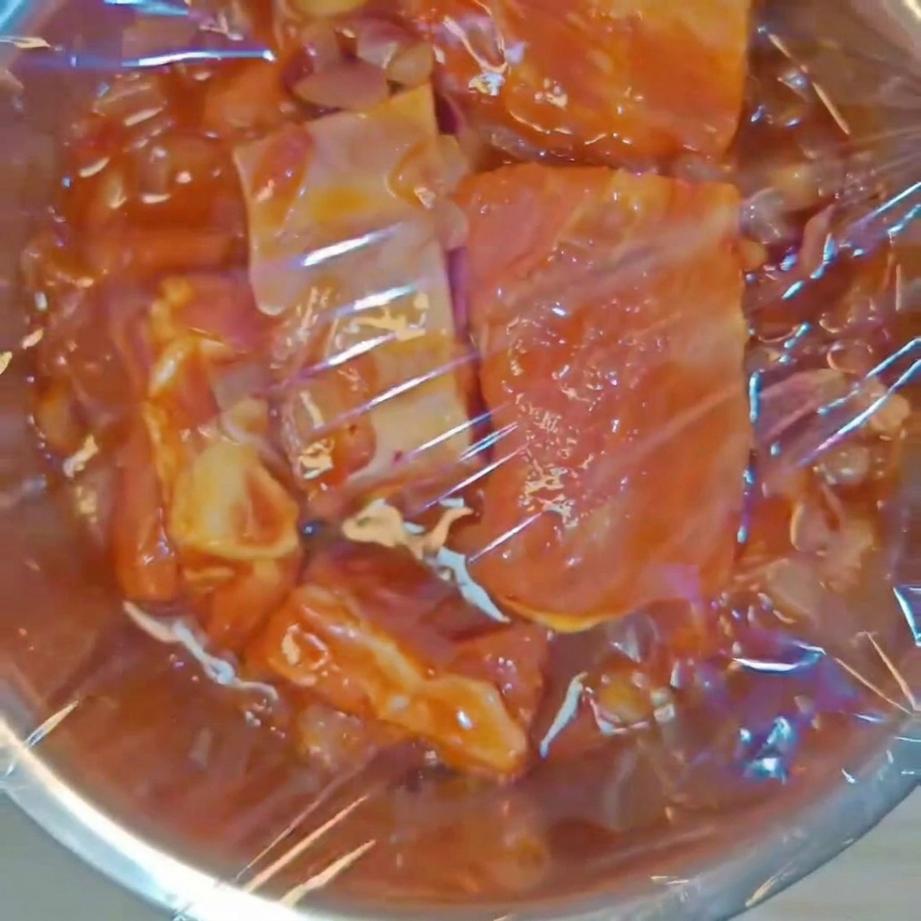 Un bol de metal con costillas de cerdo sazonadas sellado herméticamente con papel film transparente.