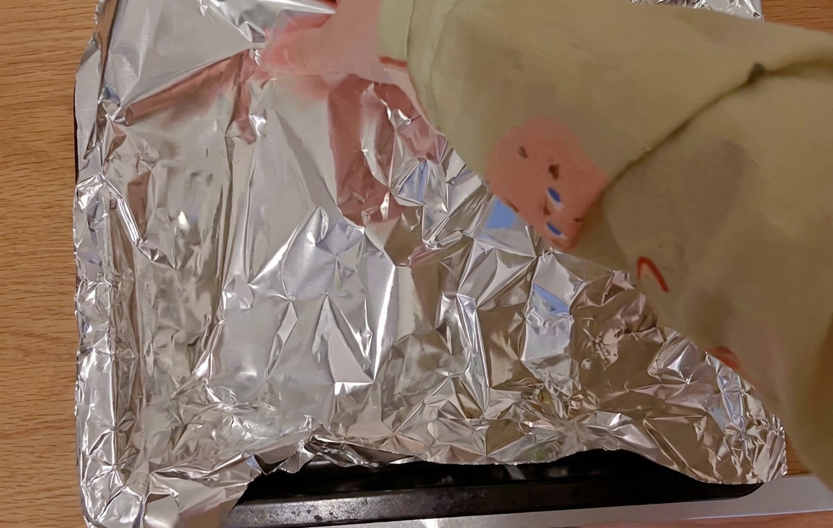 Manos cubriendo costillas de cerdo marinadas en una bandeja de horno negra con una lámina de papel de aluminio brillante.