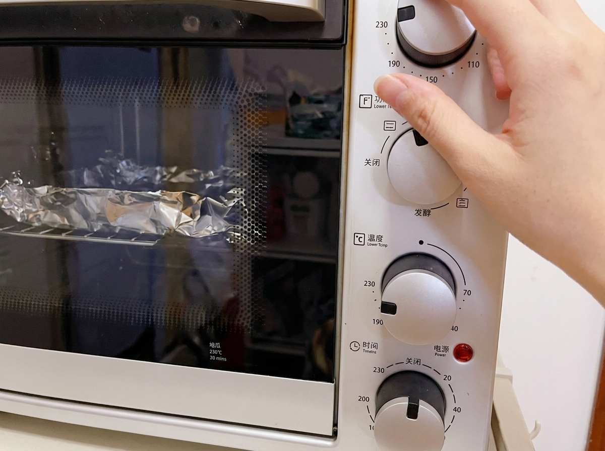 Primer plano de una mano ajustando el dial de temperatura de un horno eléctrico blanco a 230°C, con paquetes envueltos en aluminio visibles en la rejilla interior.