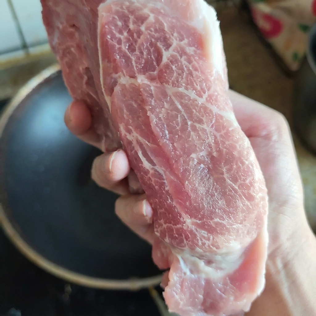 Un trozo crudo de aguja de cerdo bien veteada sostenido con la mano para mostrar su textura fresca y distribución de grasa.