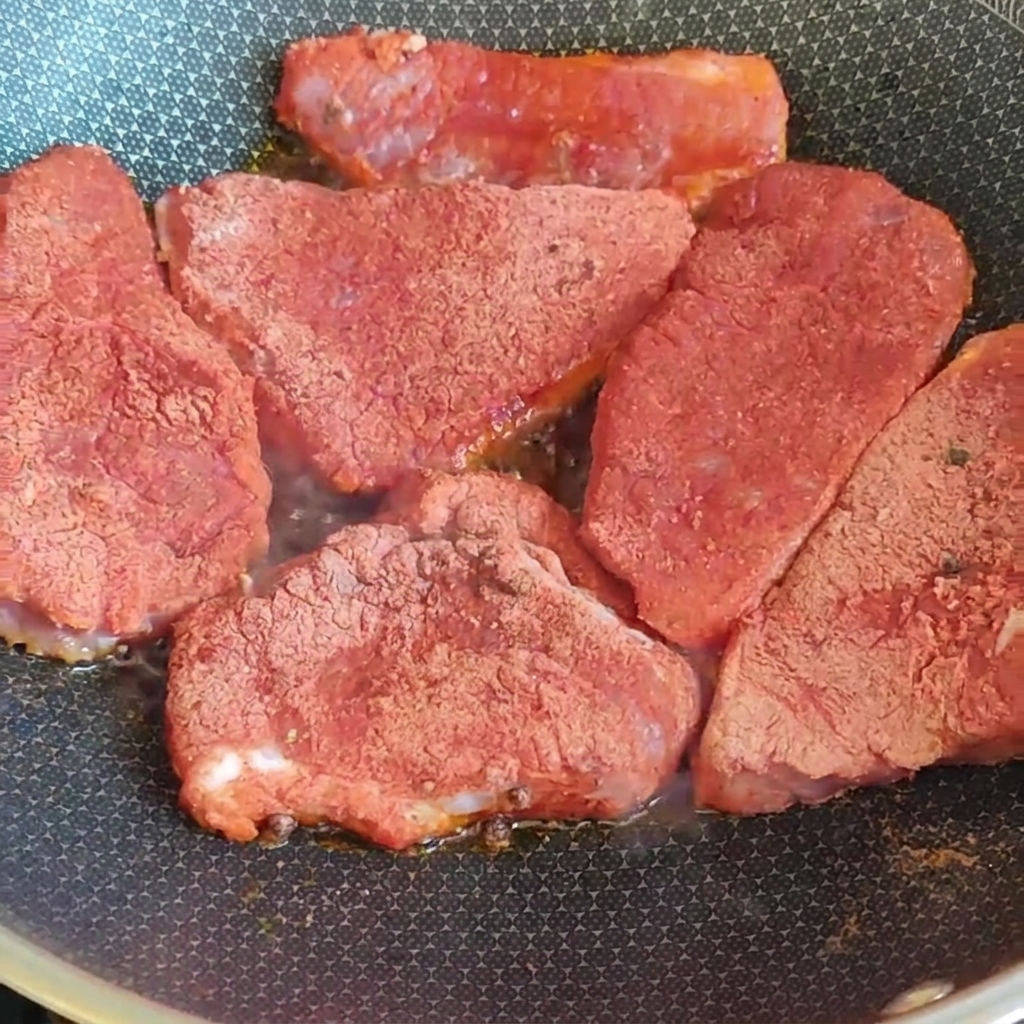 Filetes gruesos de cerdo cubiertos con una mezcla de especias rojas sellándose en una sartén caliente sobre la estufa.