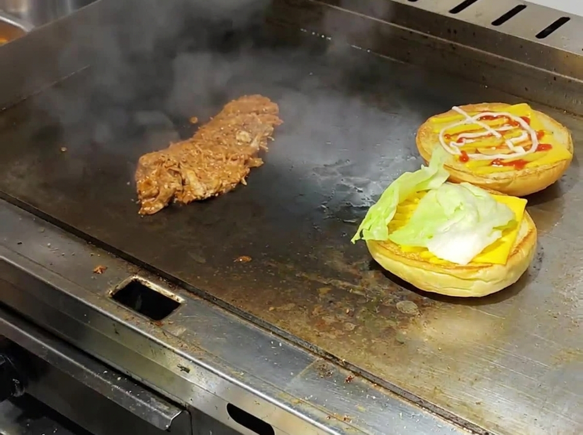 Panes de hamburguesa tostados con capas de queso y lechuga junto a carne desmechada caliente sobre una plancha comercial humeante.
