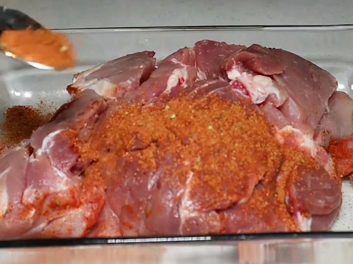 Una cucharada de especias de color rojizo esparcida sobre un trozo grande de paleta de cerdo cruda en una fuente de vidrio.