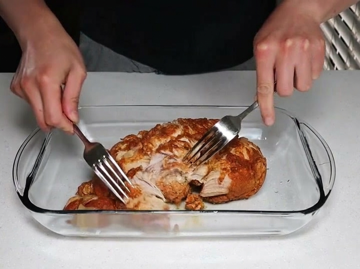 Dos manos usando tenedores de acero inoxidable para desmechar un trozo grande de carne de cerdo cocida y tierna en una fuente de vidrio.