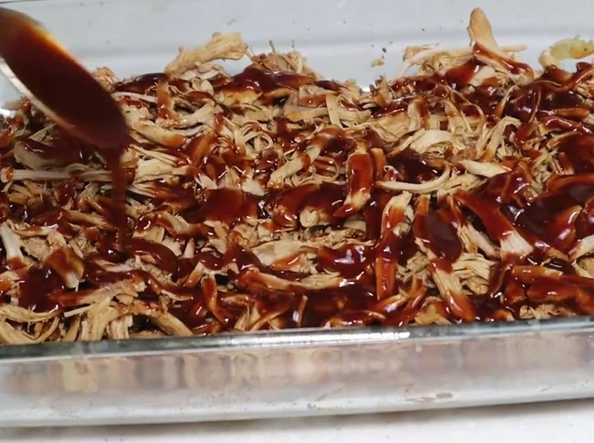 Una cucharada de salsa barbacoa espesa y oscura vertida sobre una capa de cerdo desmechado ahumado en una fuente de vidrio.