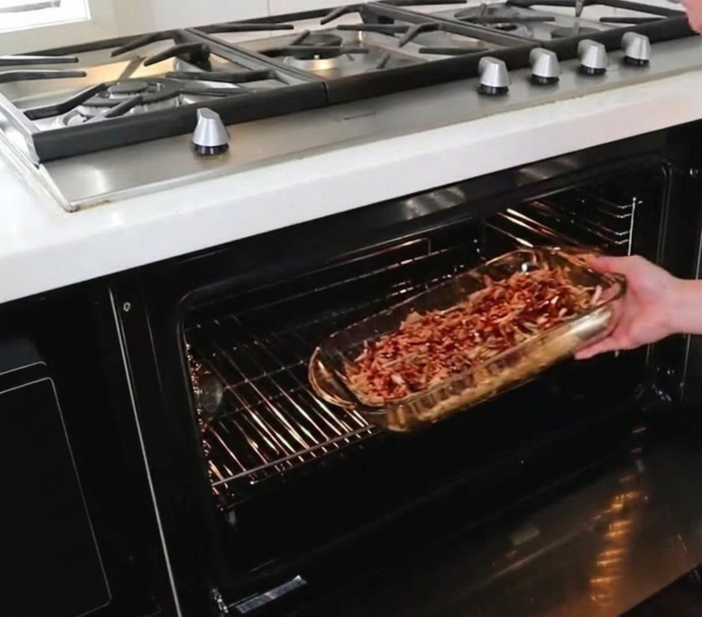 Una persona deslizando una fuente de vidrio llena de pulled pork con salsa en la rejilla central de un horno caliente.