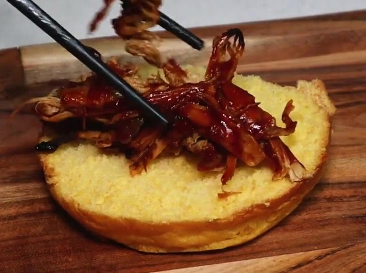 Un par de palillos colocando jugoso cerdo desmechado con salsa barbacoa sobre la superficie dorada de la mitad de un pan de hamburguesa tostado.