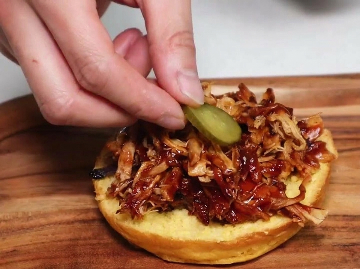 Una mano colocando una rodaja redonda de pepinillo sobre una porción de pulled pork con salsa en un pan de hamburguesa tostado.