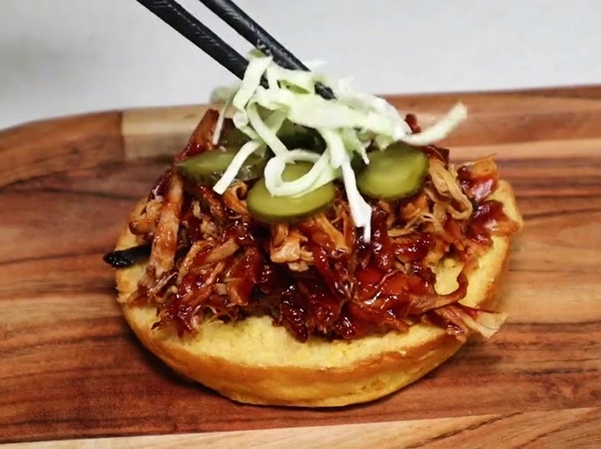 Unos palillos colocando ensalada de col blanca rallada sobre las rodajas de pepinillo y el pulled pork en un pan de hamburguesa.