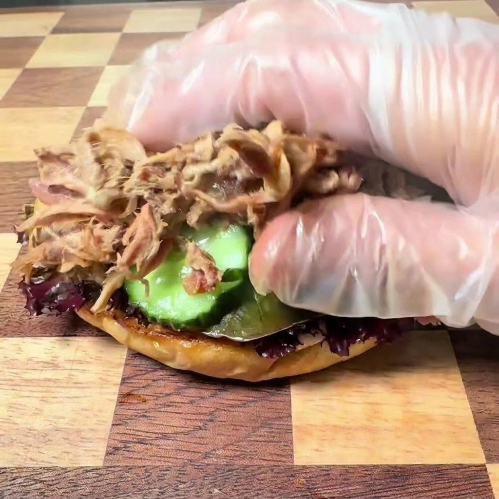Una mano con guante de plástico transparente colocando tierno pulled pork deshebrado sobre la base de la hamburguesa con lechuga y pepinillos.