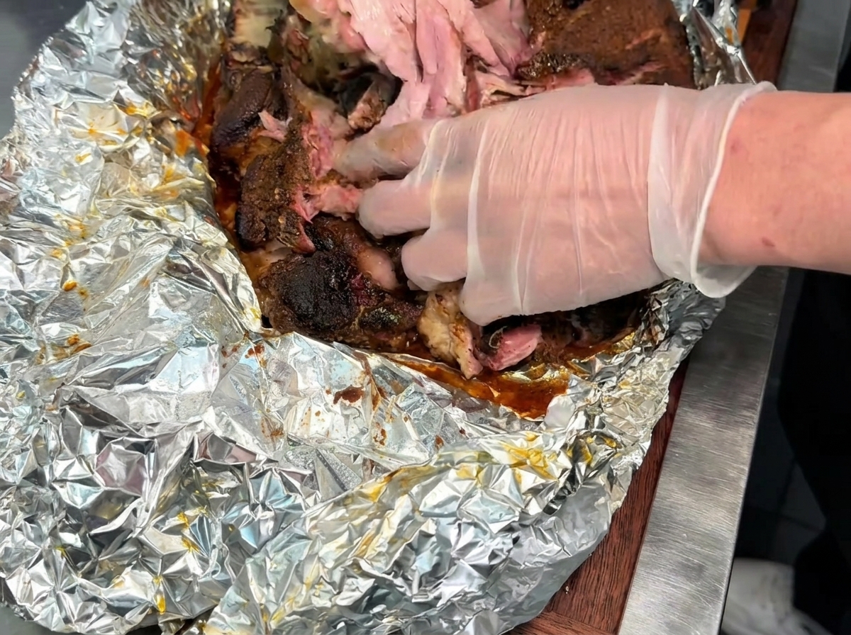 Manos con guantes desmenuzando y deshebrando una pieza grande de carne de cerdo ahumada y tierna sobre una superficie cubierta con papel de aluminio.