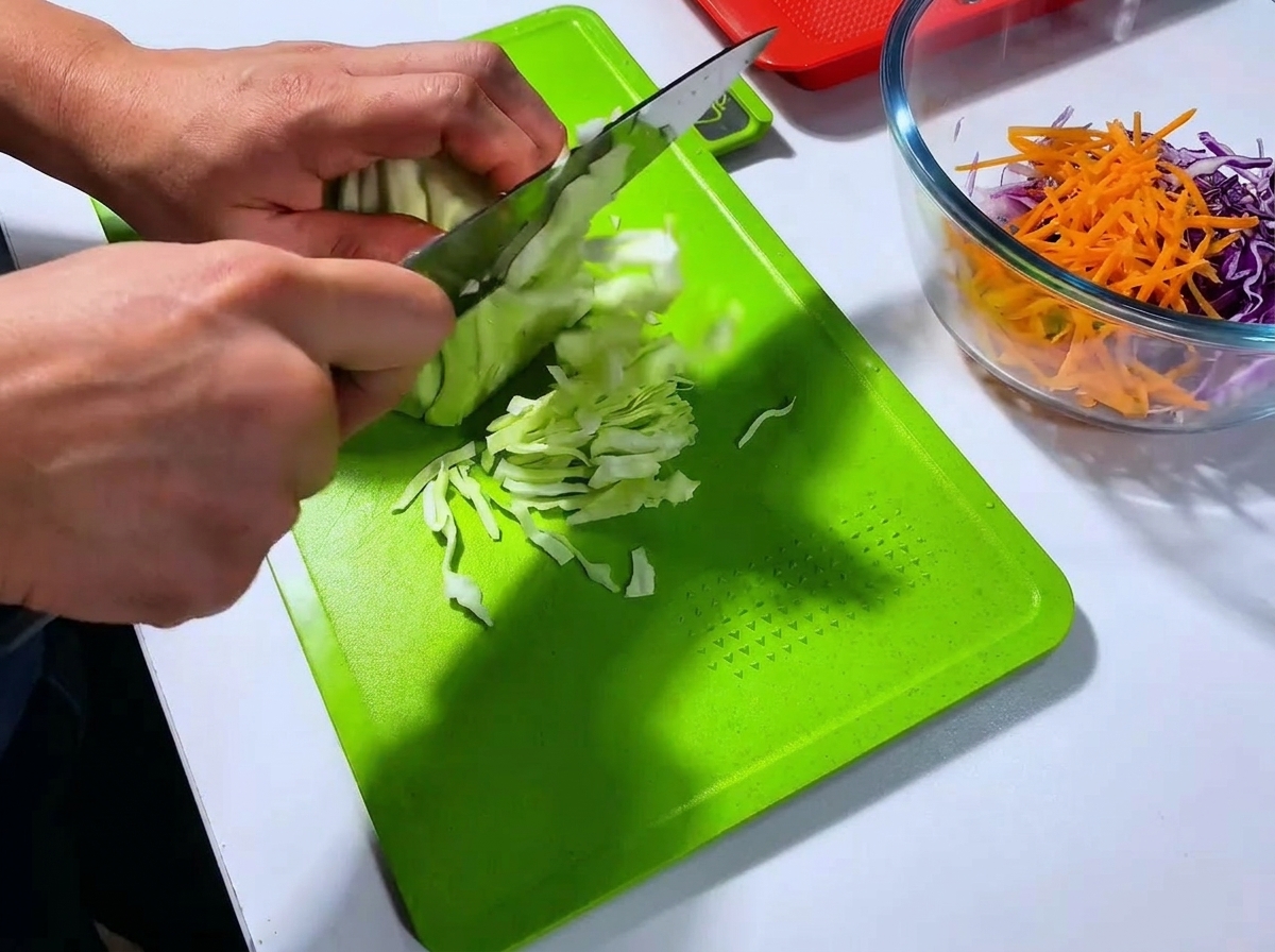 Un chef cortando una cabeza de col blanca en tiras finas con un cuchillo grande sobre una tabla de cortar de color verde brillante.