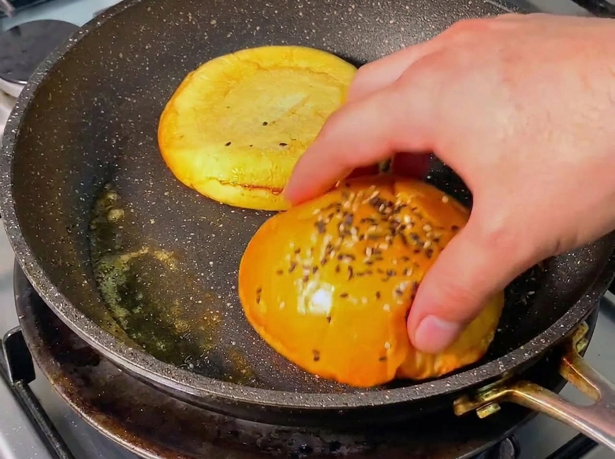 Una mano dando la vuelta a la mitad de un pan de hamburguesa dorado en una sartén caliente con mantequilla derretida.