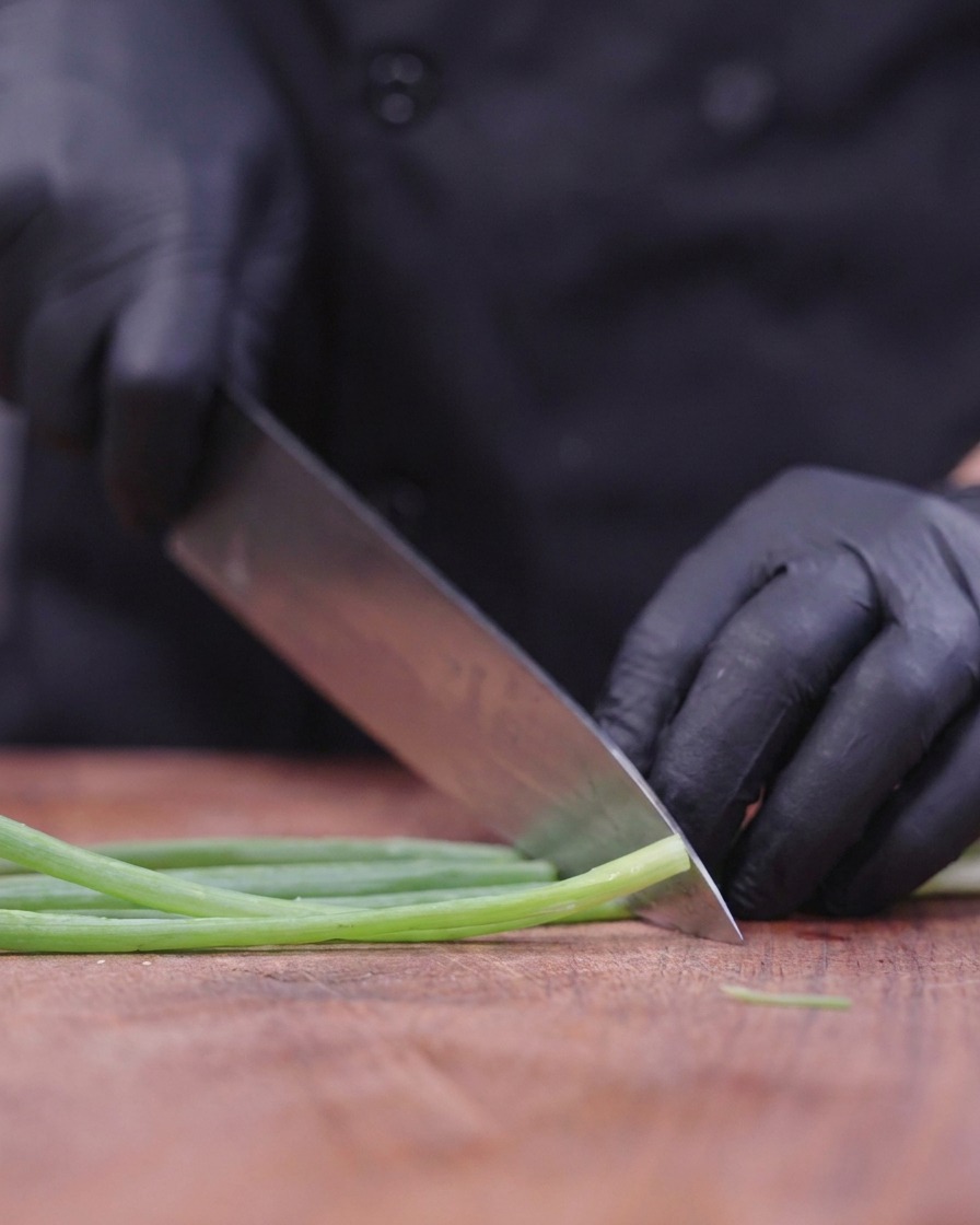 Un chef con guantes negros utilizando un cuchillo grande para picar finamente tallos de cebolleta en una tabla de madera.