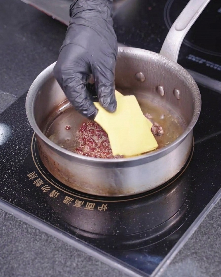 Una mano con guante colocando una loncha cuadrada de queso amarillo sobre la carne de hamburguesa en una sartén caliente.