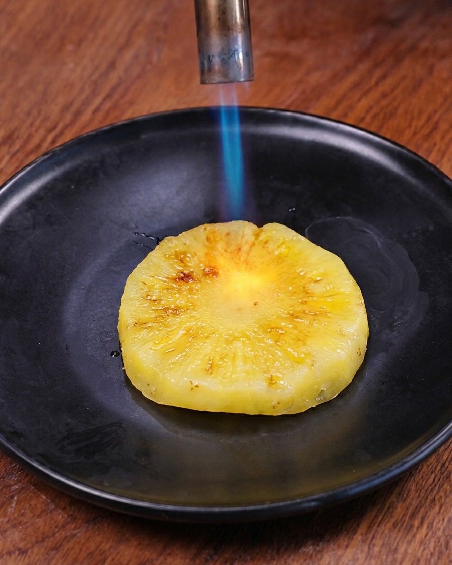 Una llama azul de un soplete de cocina quemando la superficie de un disco de piña fresca en un plato oscuro.