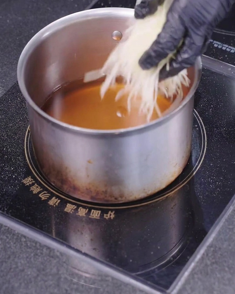 Una mano con un guante negro dejando caer patatas cortadas muy finas en un cazo lleno de aceite caliente.