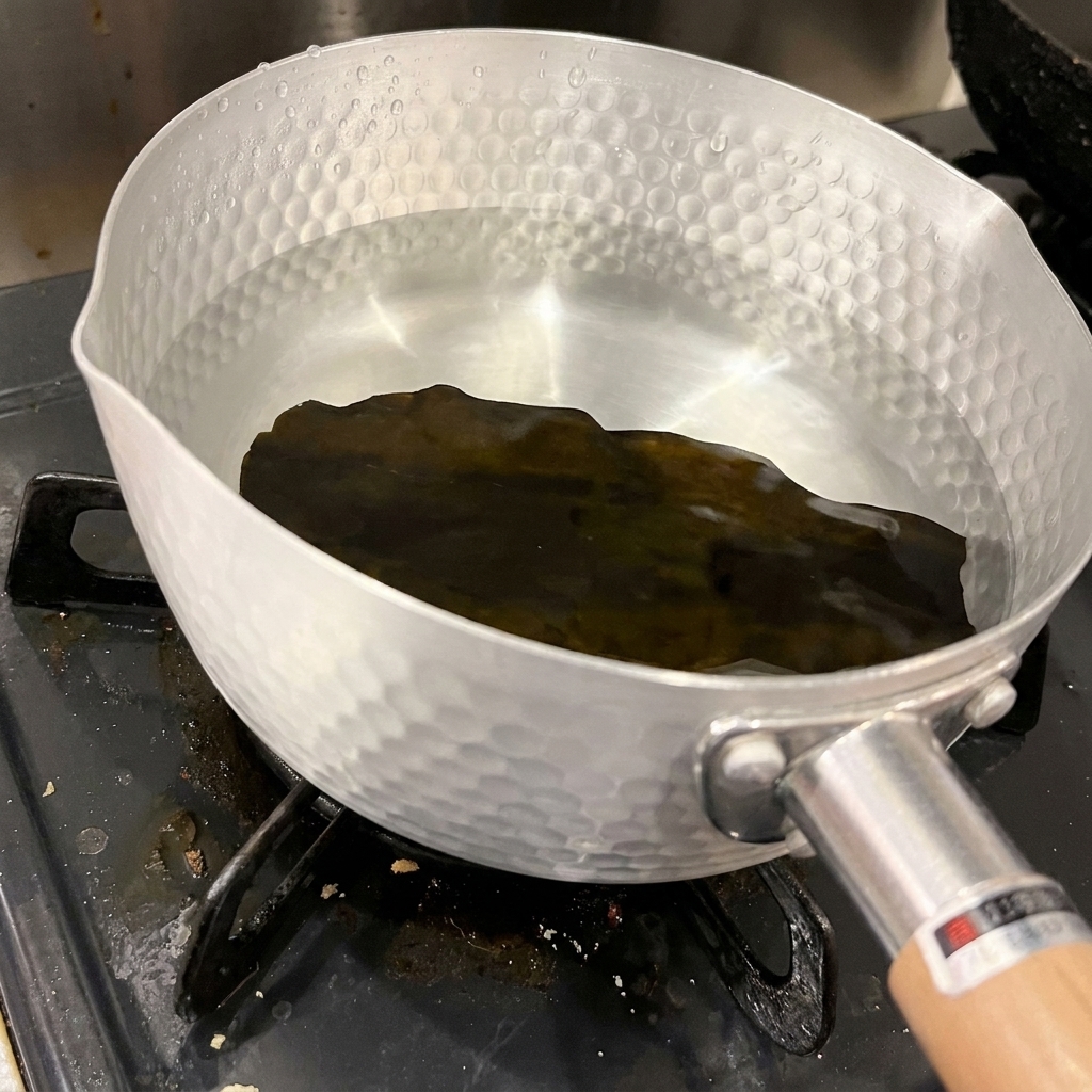 Un trozo de alga kombu seca y oscura en el fondo de una olla de aluminio con agua fría y transparente.
