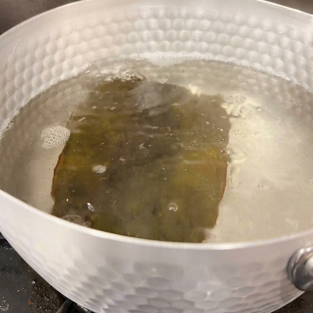 Agua burbujeando suavemente alrededor de un trozo de alga kombu rehidratada dentro de una olla plateada.