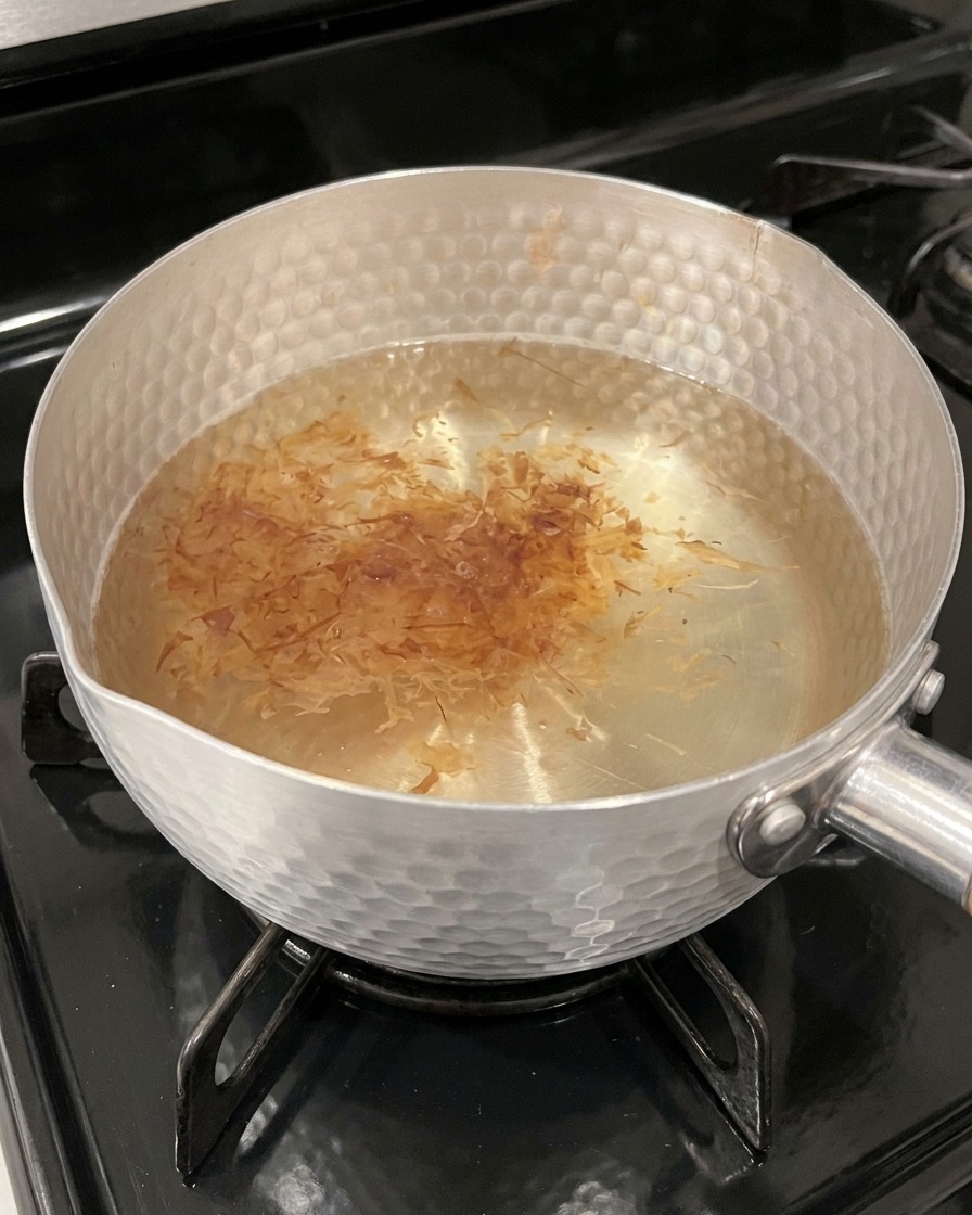 Copos de bonito ligeros y secos flotando en la superficie de un caldo caliente de tono ámbar en una olla.