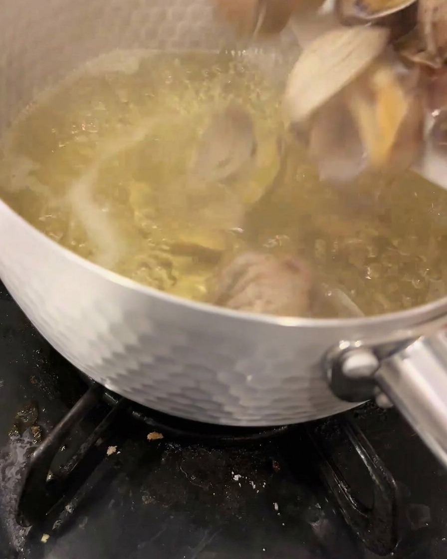 Almejas frescas introduciéndose en un caldo dashi hirviendo dentro de una olla metálica sobre un fuego de gas.