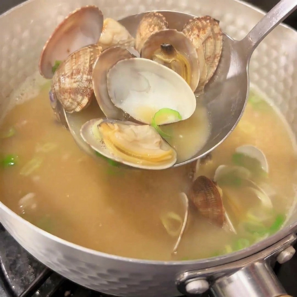 Un cazo de metal sirviendo almejas abiertas de una olla de sopa de miso japonesa humeante con cebollino picado brillante.