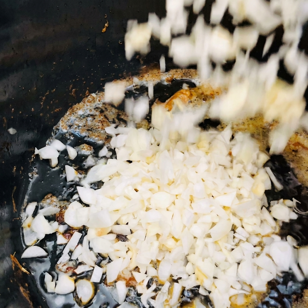 Cebollas blancas picadas y ajo picado cayendo en una olla oscura con aceite caliente brillante.
