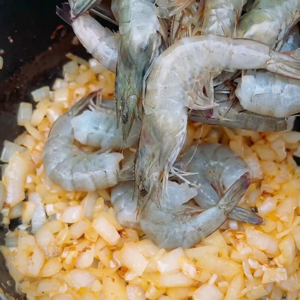 Camarones enteros frescos de color gris colocados sobre una cama de cebollas picadas sofritas en una olla.