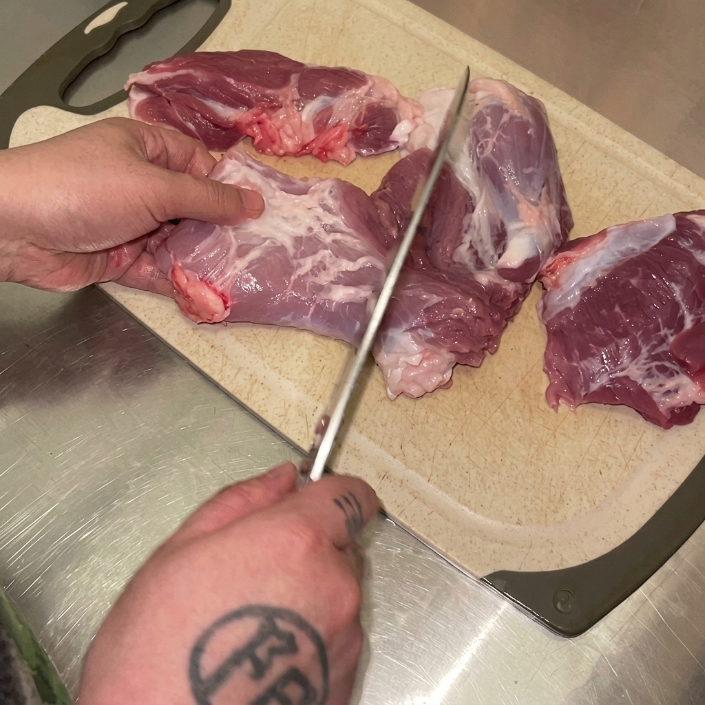 Manos usando un cuchillo para cortar carne cruda de pierna de cerdo en trozos sobre una tabla de cortar.