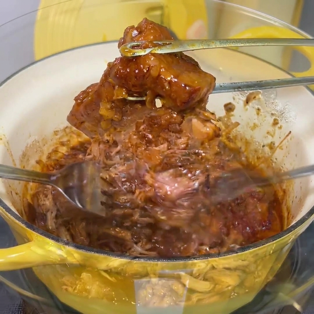 Carne de cerdo cocinada siendo desmechada en hilos finos con dos tenedores de metal en un bol de cristal.