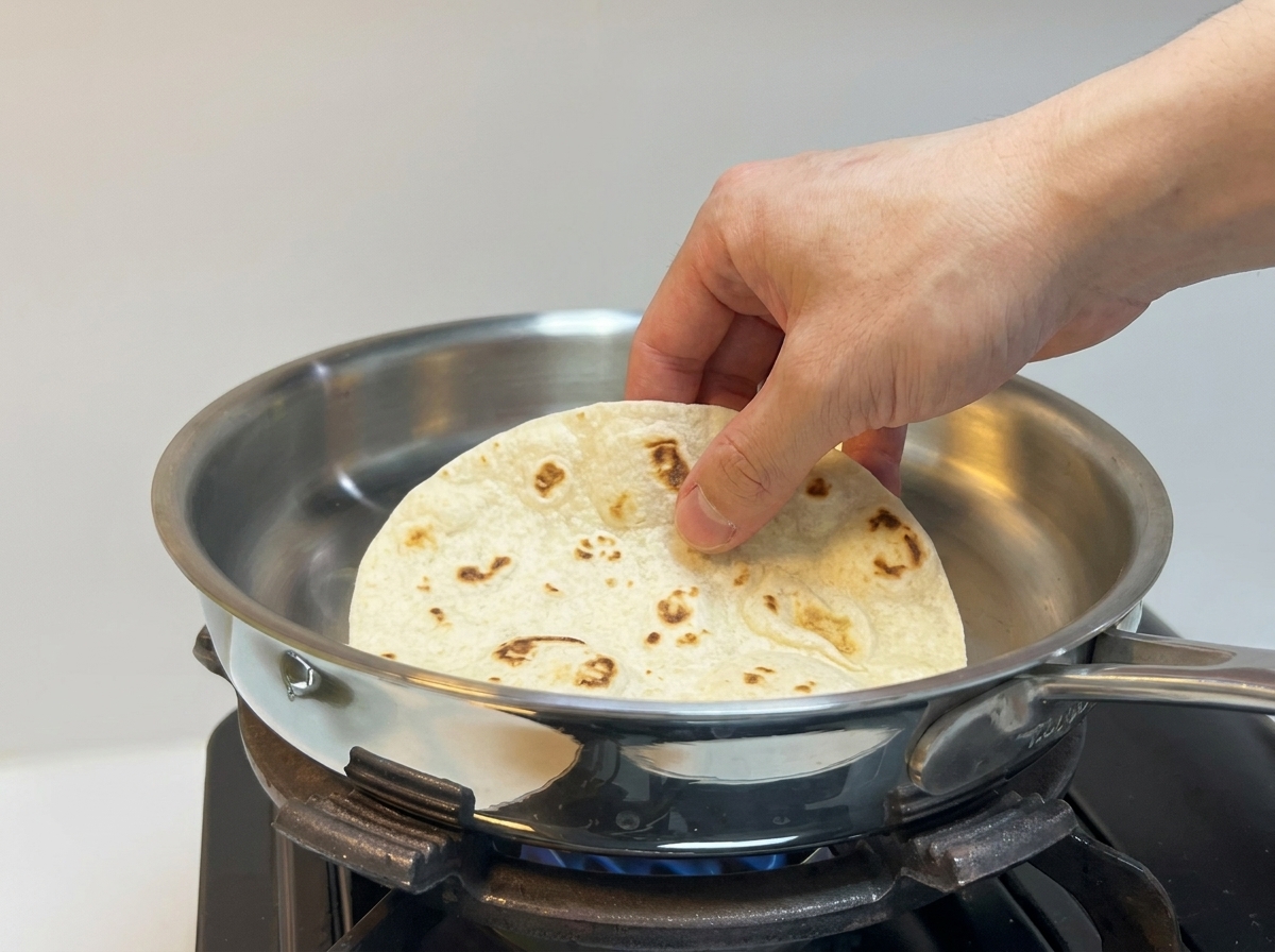 Una mano colocando una tortilla de harina gruesa sobre una sartén de acero inoxidable caliente.