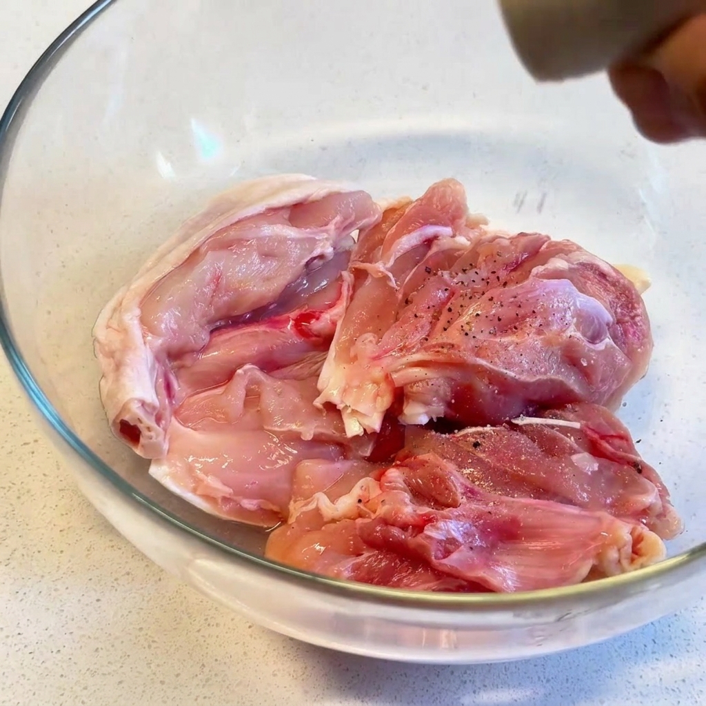 Muslos de pollo crudos en un bol de cristal transparente siendo sazonados con pimienta negra recién molida.