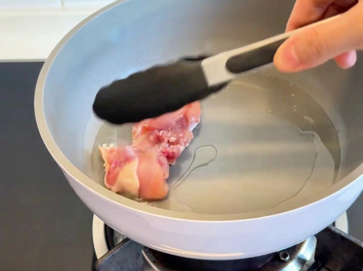 Muslos de pollo dorándose con la piel hacia abajo en una sartén caliente, mostrando una piel crujiente y dorada.