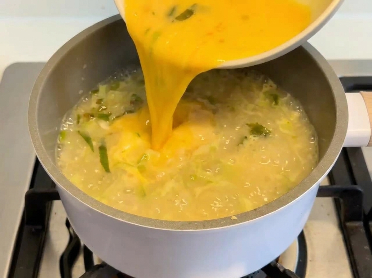 Una vibrante mezcla amarilla de huevo y limón siendo vertida desde un bol blanco a una olla humeante de sopa cremosa.