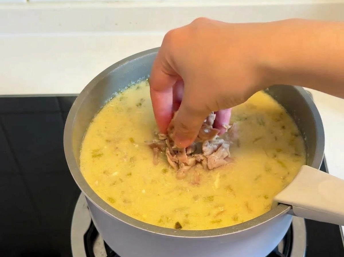 Una mano echando trozos de pollo cocido y desmenuzado en una olla con sopa de limón espesa y cremosa.