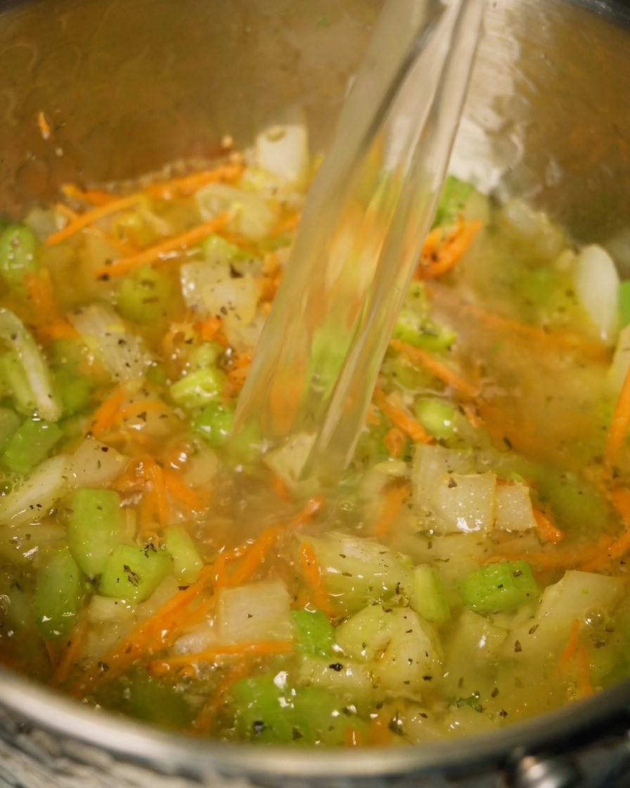 Caldo de pollo vertiéndose en una olla que contiene verduras y hierbas sofritas.