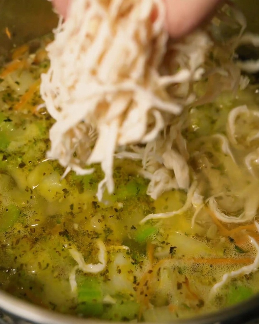 Pollo deshebrado agregándose a una olla de sopa hirviendo a fuego lento.