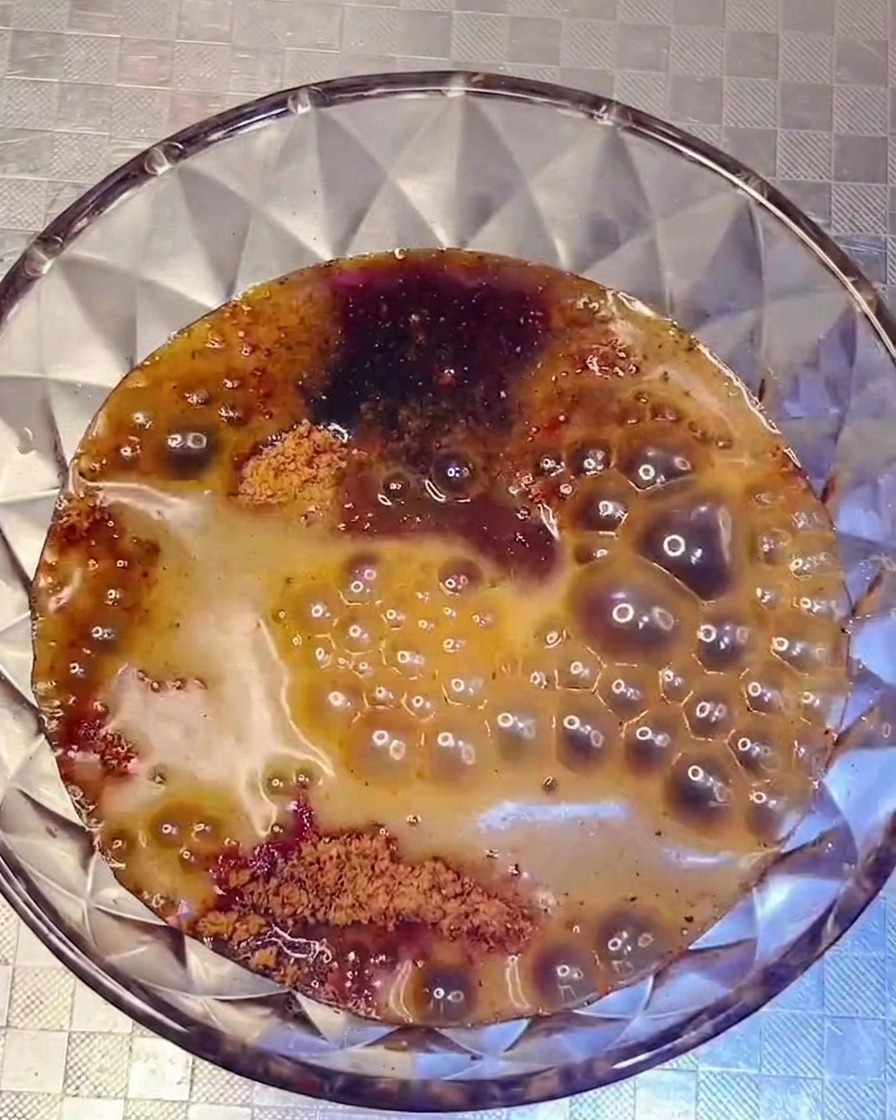 Una mezcla de salsa oscura y sabrosa siendo vertida desde un bol de vidrio hacia una olla llena con varios ingredientes organizados.