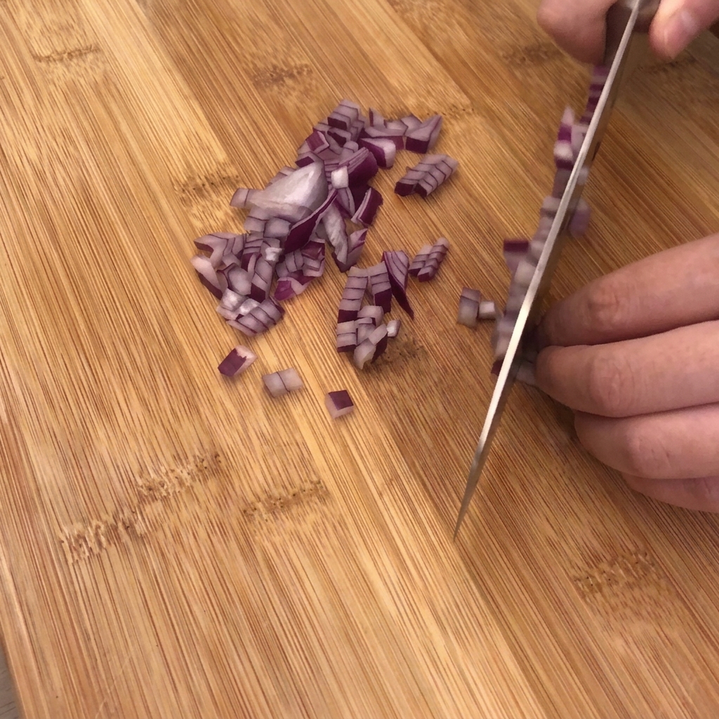 Un cuchillo afilado picando una cebolla morada en una tabla de cortar de madera con trozos de ajo y tocino cerca.